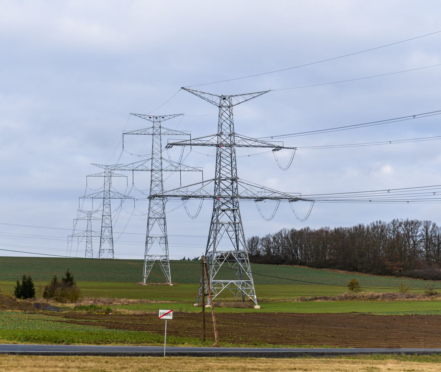 High voltage power lines stretch across a rural field beneath a cloudy sky.
