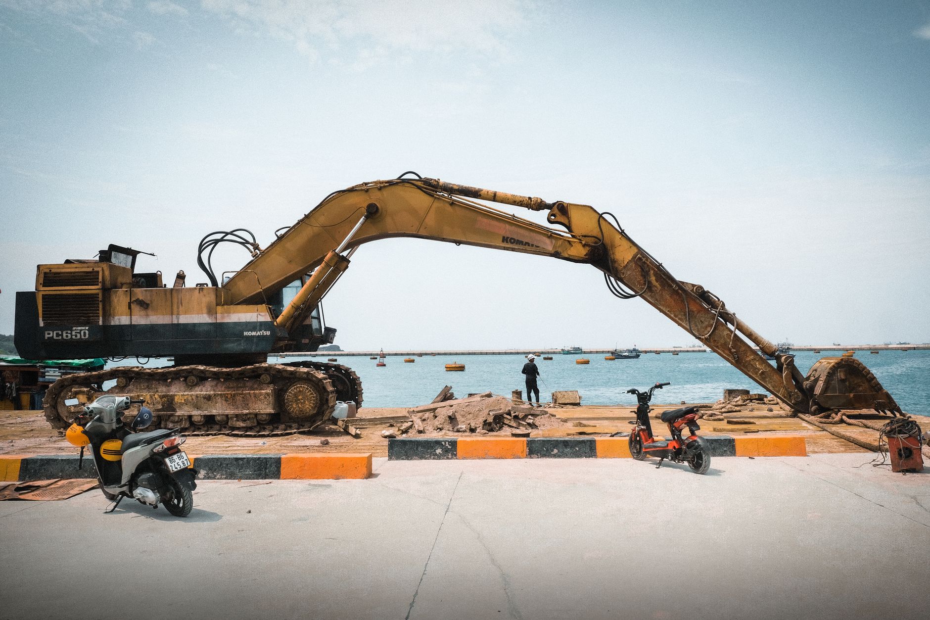 A large excavator working on a coastal construction site with a sea view.