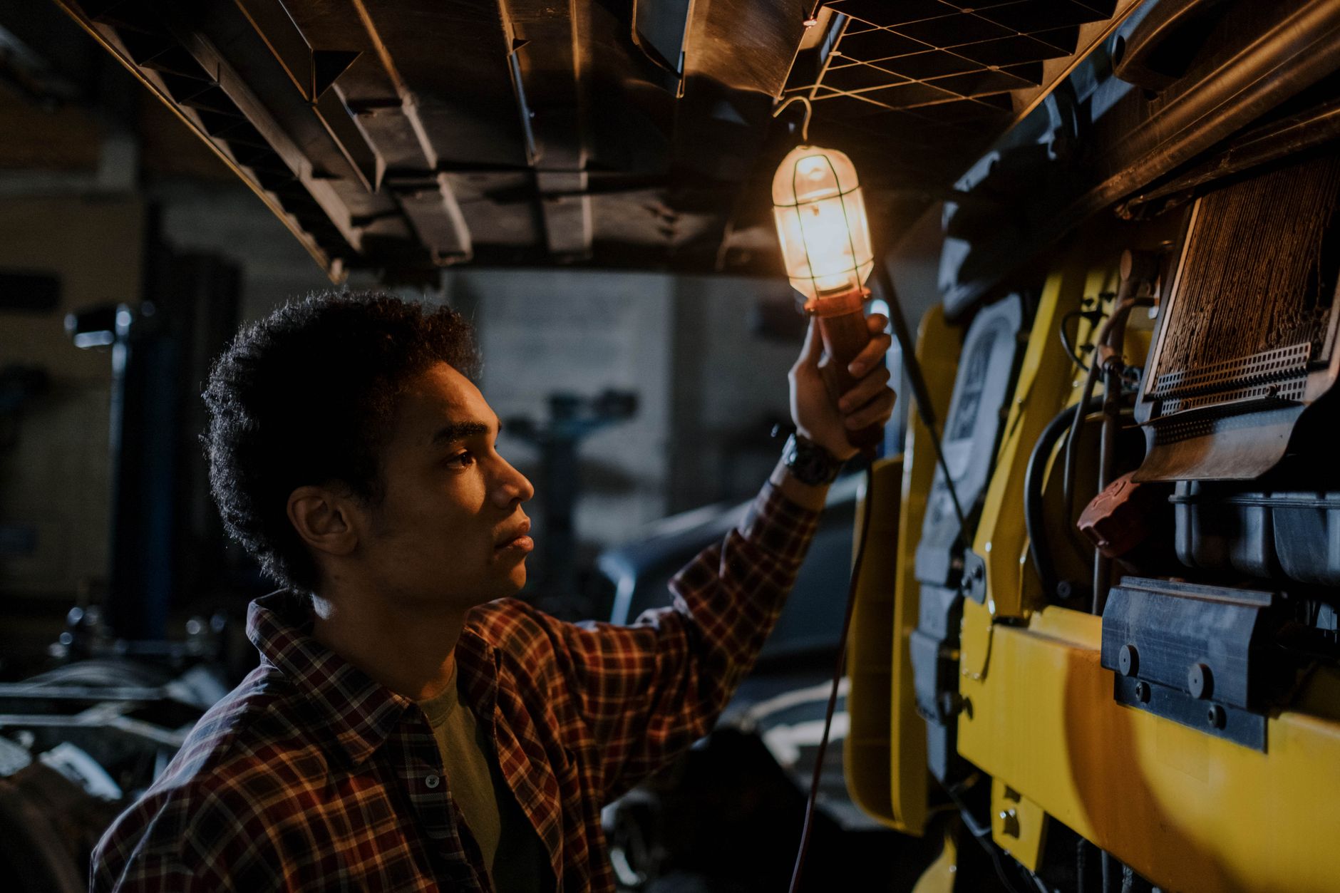 Auto mechanic inspecting a truck engine with a trouble light in a dimly lit garage.
