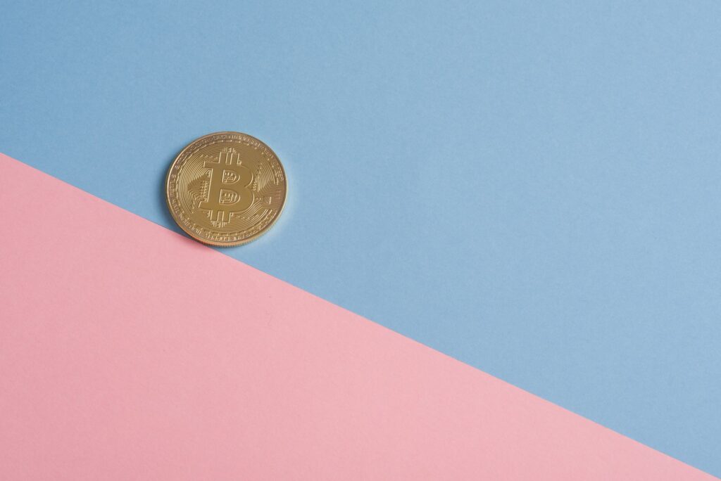 A single gold Bitcoin on a minimal pastel blue and pink background.