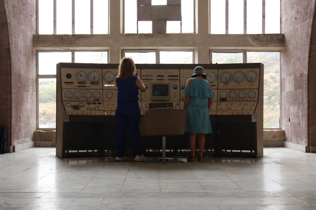 Two women operate a vintage control panel in a spacious industrial room with large windows.
