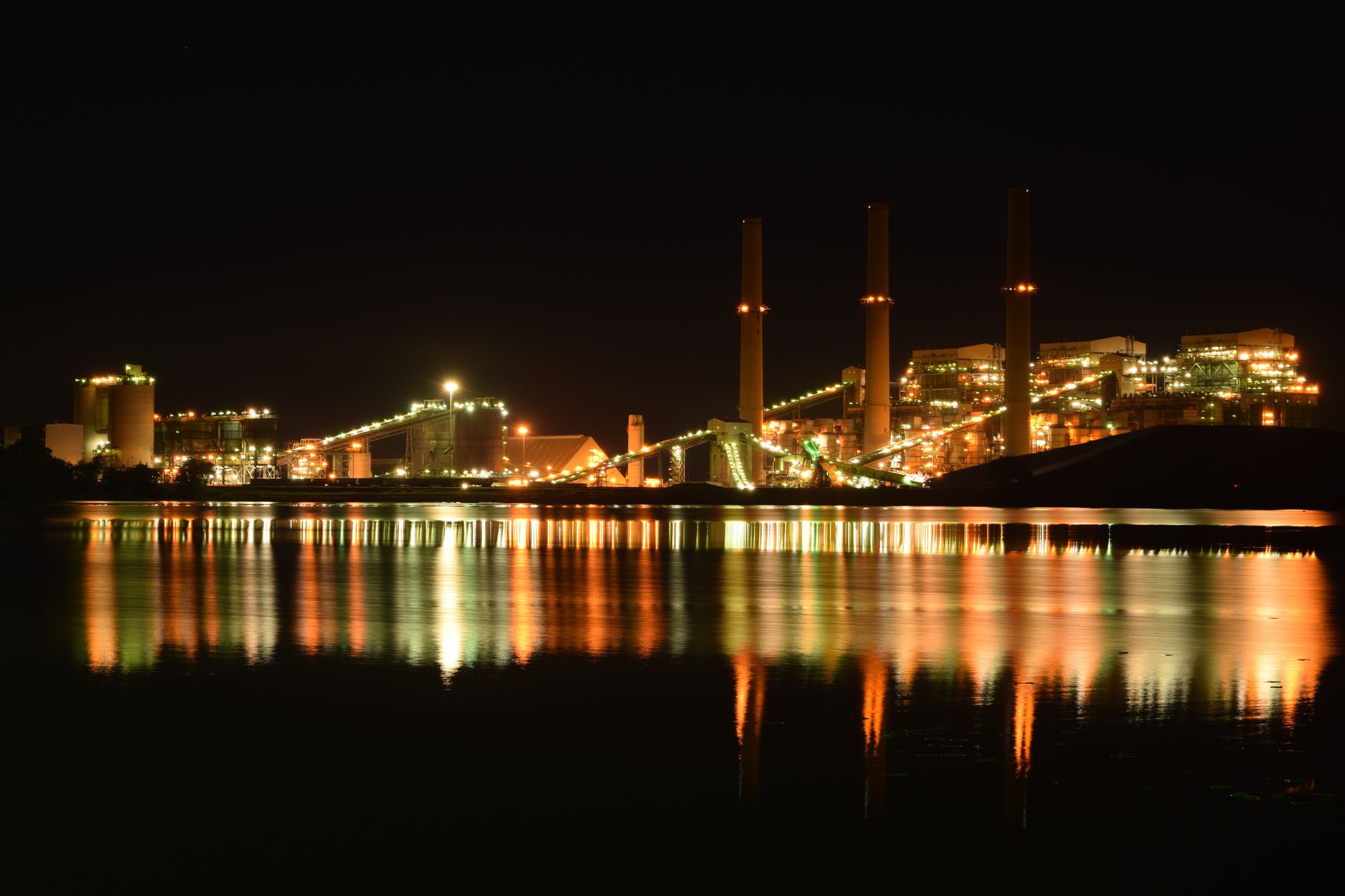 A brightly lit industrial plant reflected in a river at night creates a dramatic scene.