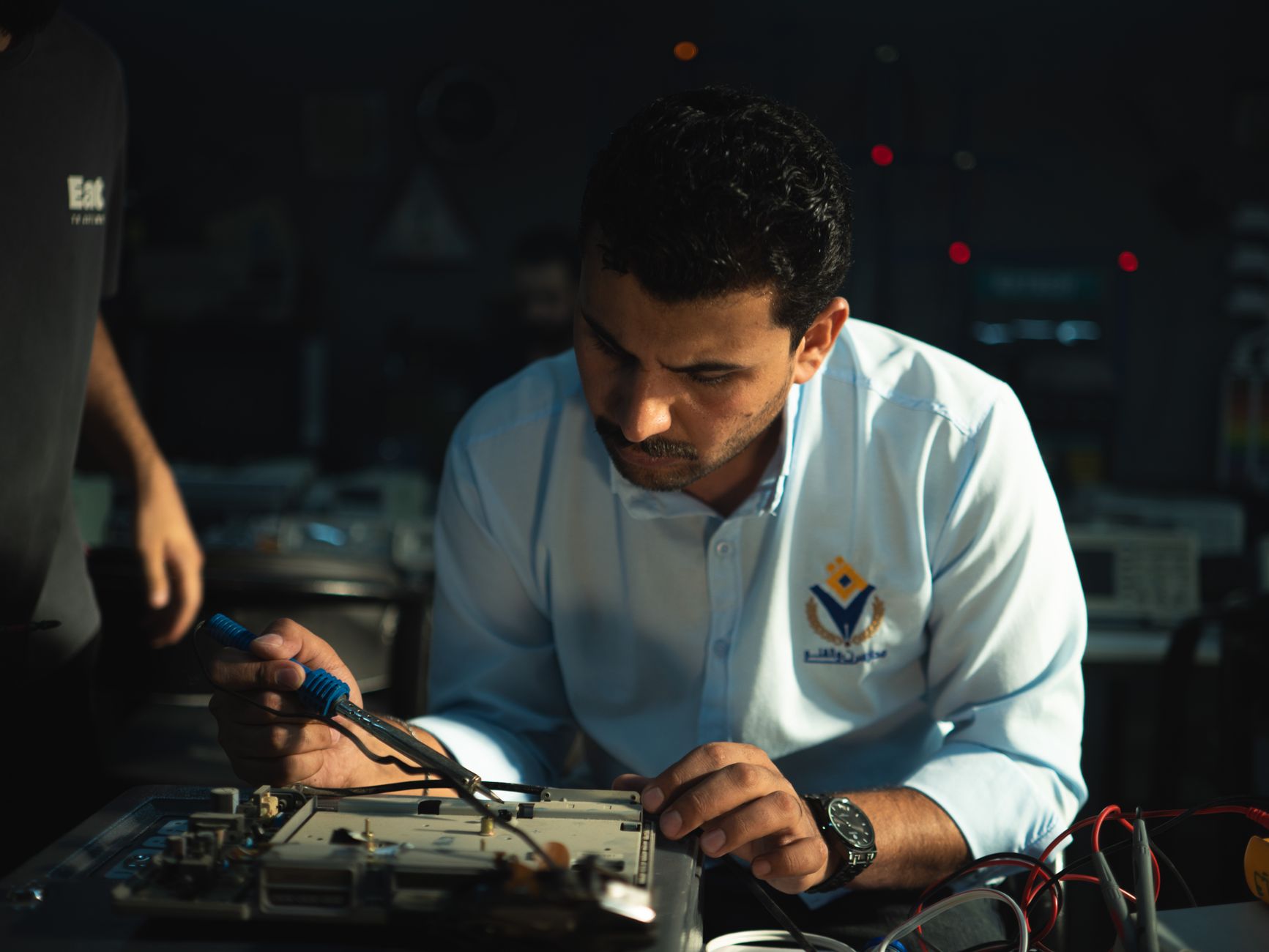 Middle Eastern technician soldering electronic components at a workshop bench with focused attention.