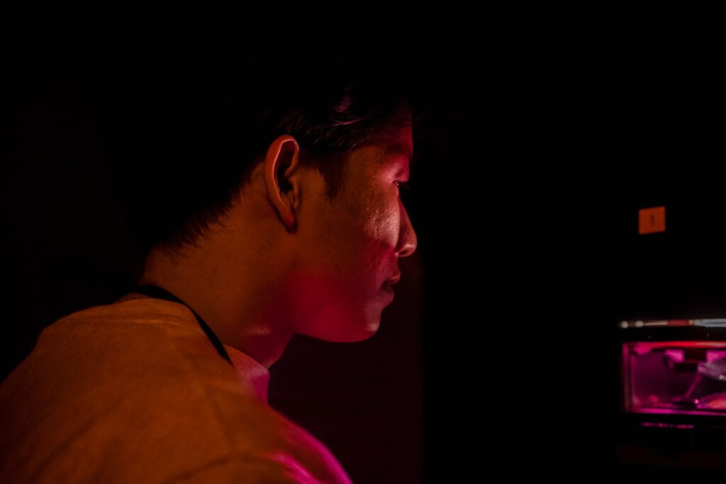 Side profile of an Asian man in a pink spotlight against a dark indoor setting.